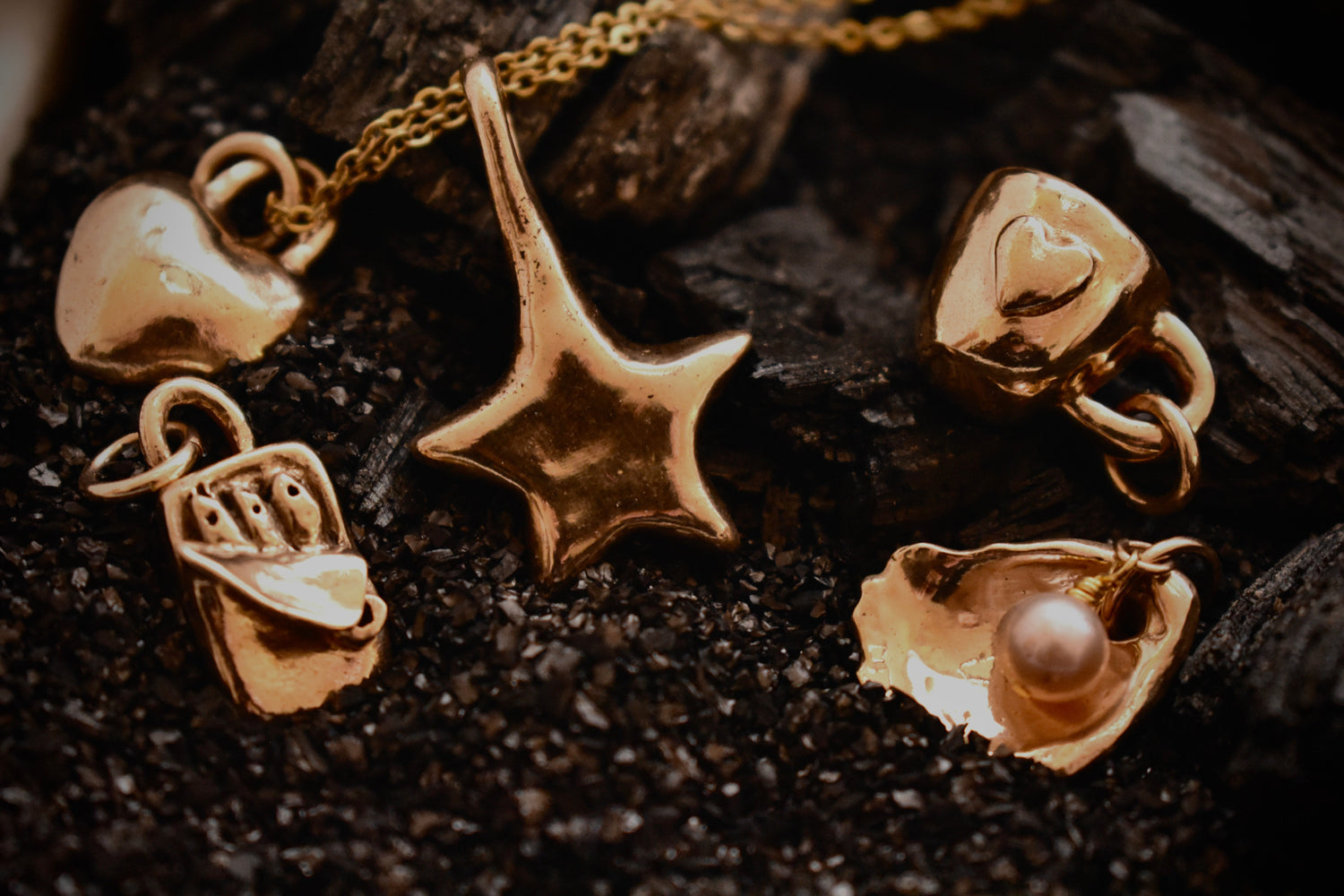 bronze charms displayed on a gold chain. From left to right: Bronze sardine tin, bronze coffee mug, bronze star, bronze oyster with pink pearl, bronze heart. All hand-sculpted from bronze clay. Laying against a blue velvet background.